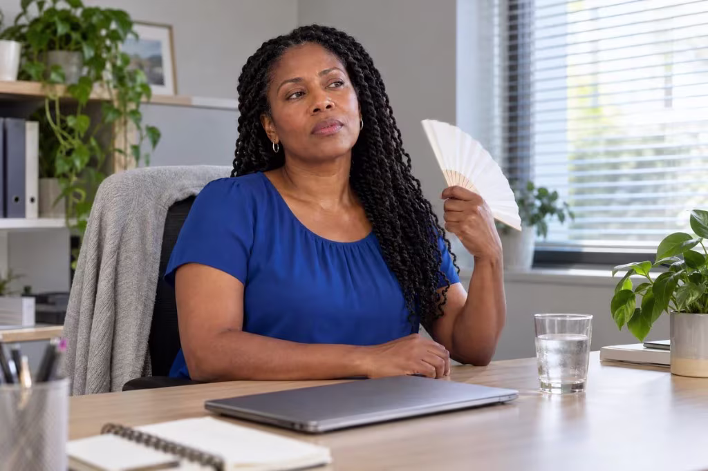 Hot flashes in perimenopause: a woman cools herself with a hand fan at her desk.