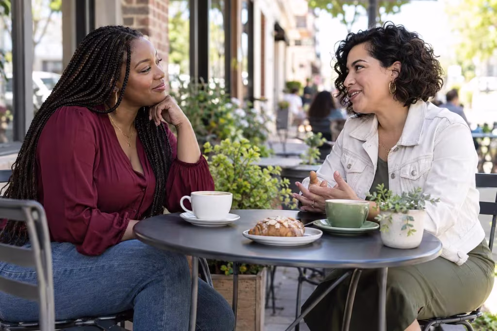 Two women talking over coffee at a small outdoor cafe table, one listening with her chin on her hand