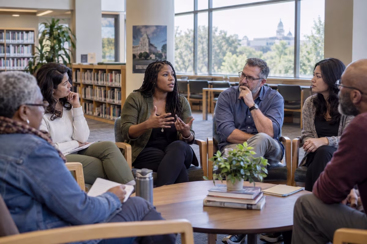A diverse group sits in a circle in a bright library, listening as one woman speaks and gestures.