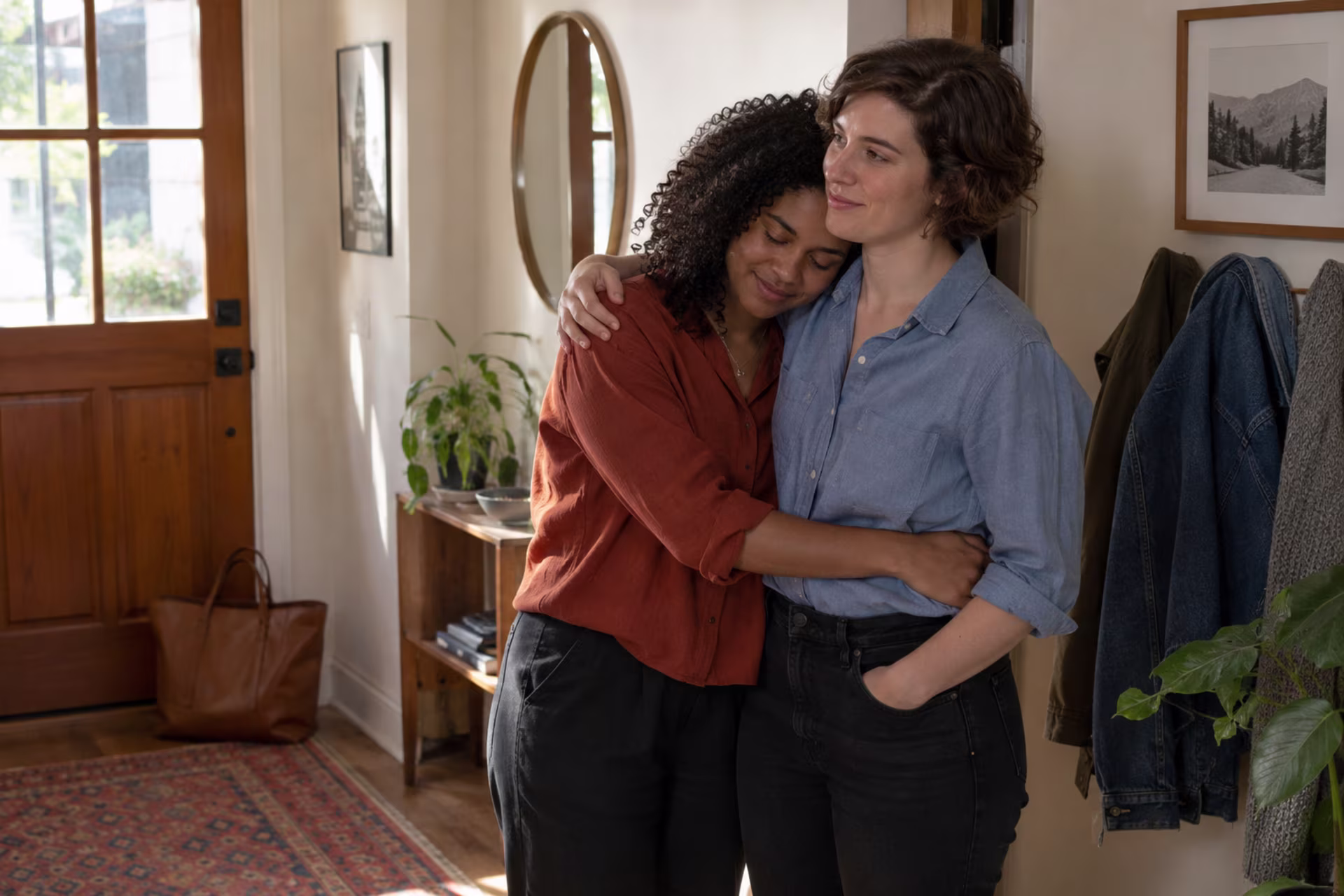Two women hug in a sunlit hallway near a wooden front door, coats hanging on the wall.
