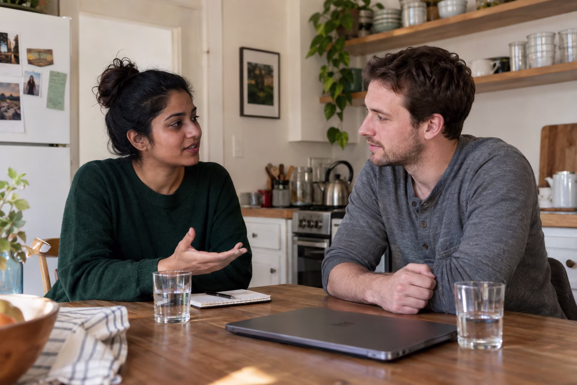 A woman and a man sit at a wooden kitchen table with water glasses and a closed laptop, talking.