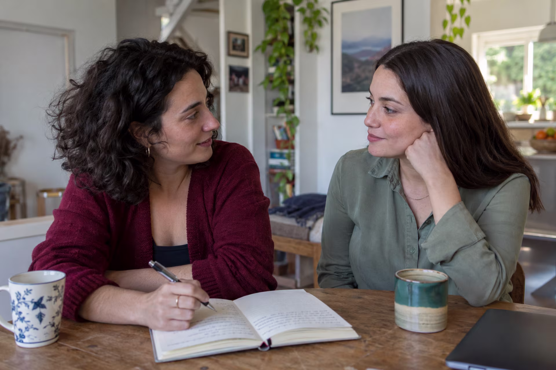 Two women sit at a wooden table with coffee mugs while one writes in an open notebook and they talk.