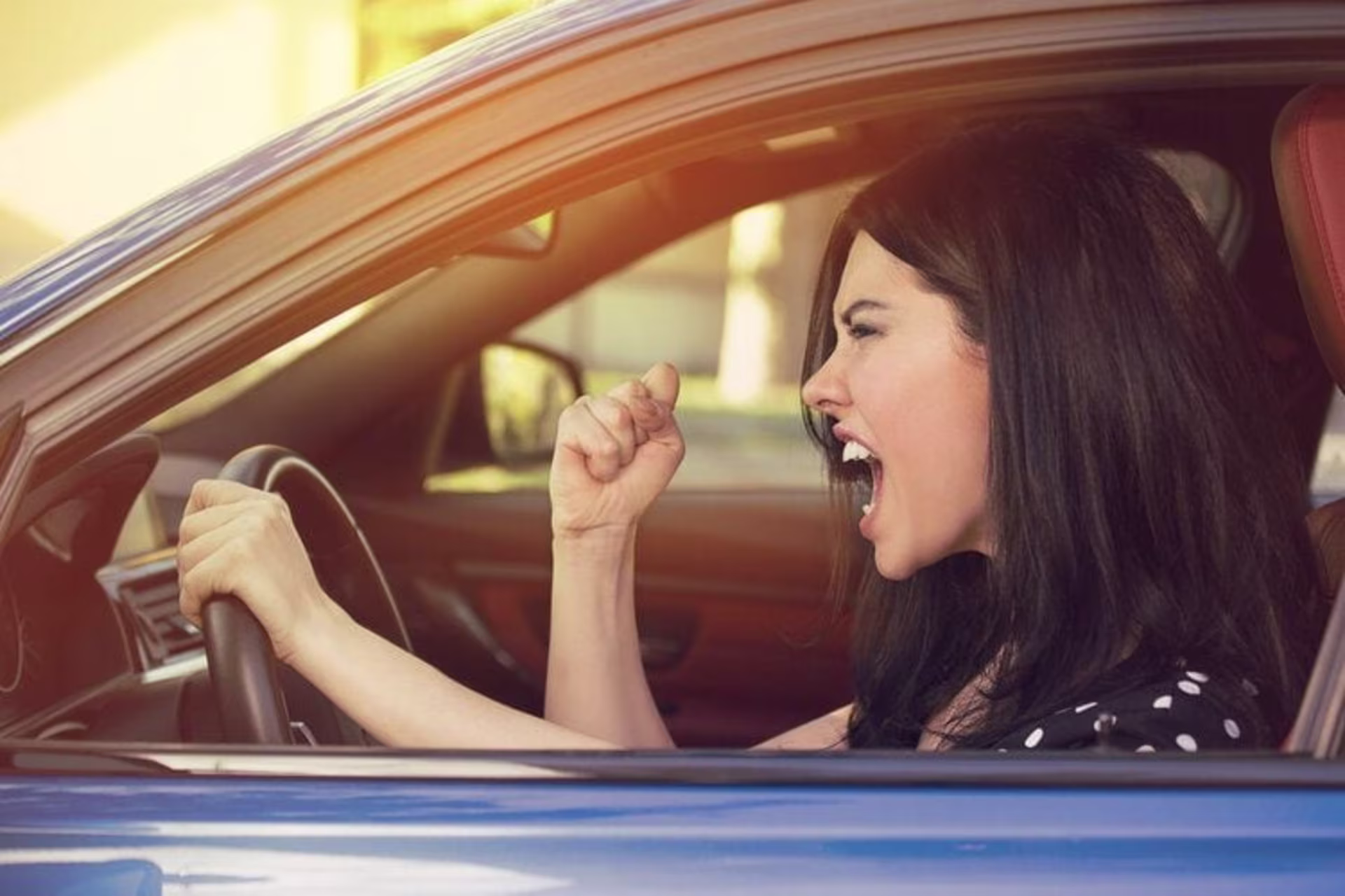 A woman in a blue car shouts and raises her fist while gripping the steering wheel with her other hand, clearly in need 