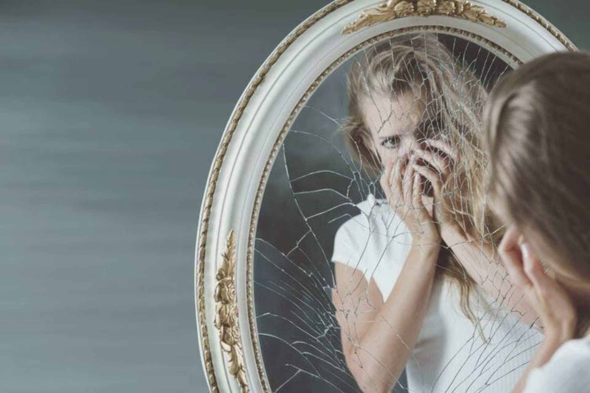 A woman wearing a white shirt stands in front of a cracked oval mirror, covering her mouth with her hands as she looks a