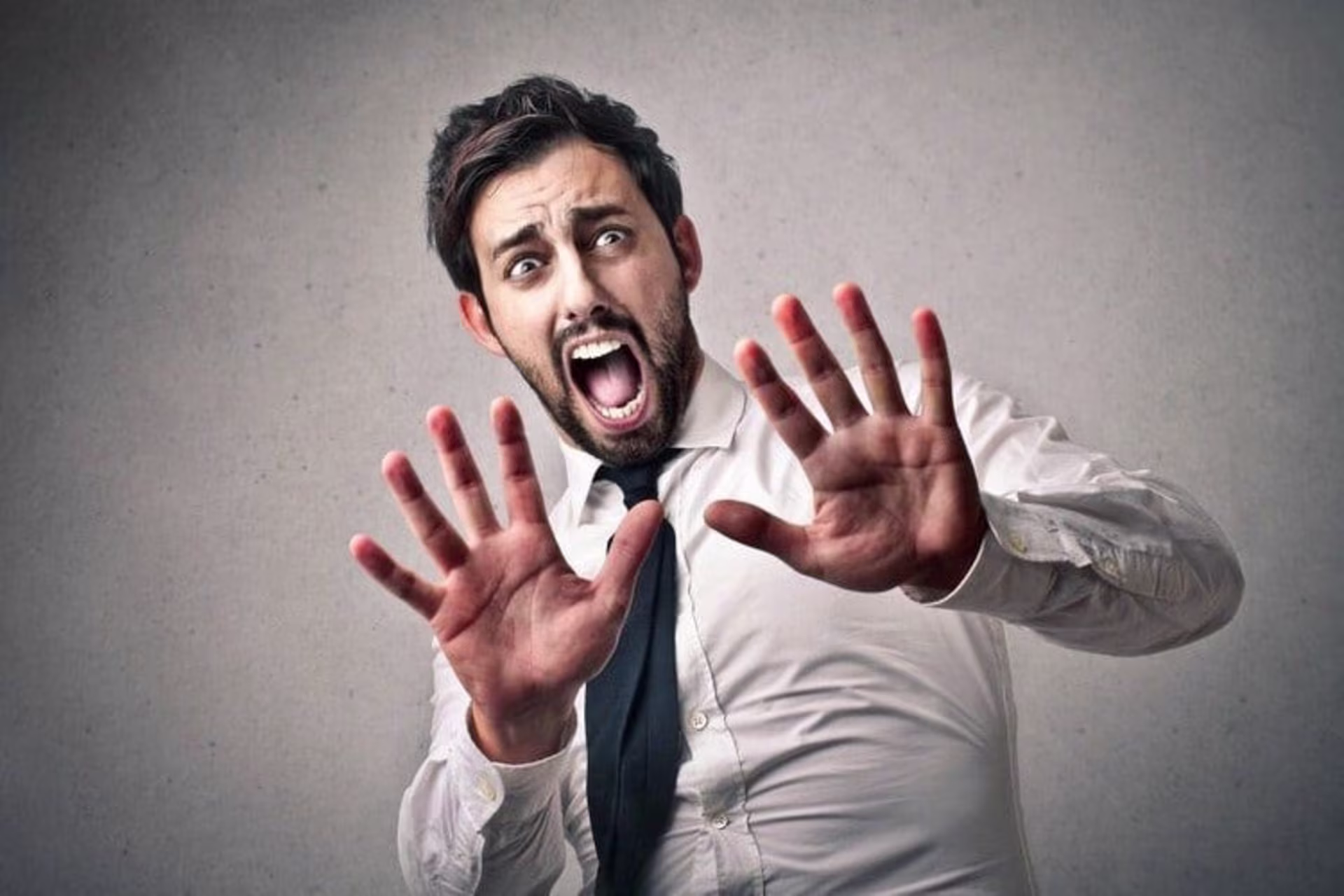 A man in a white shirt and tie is captured with a frightened expression, holding up both hands in a defensive gesture. I