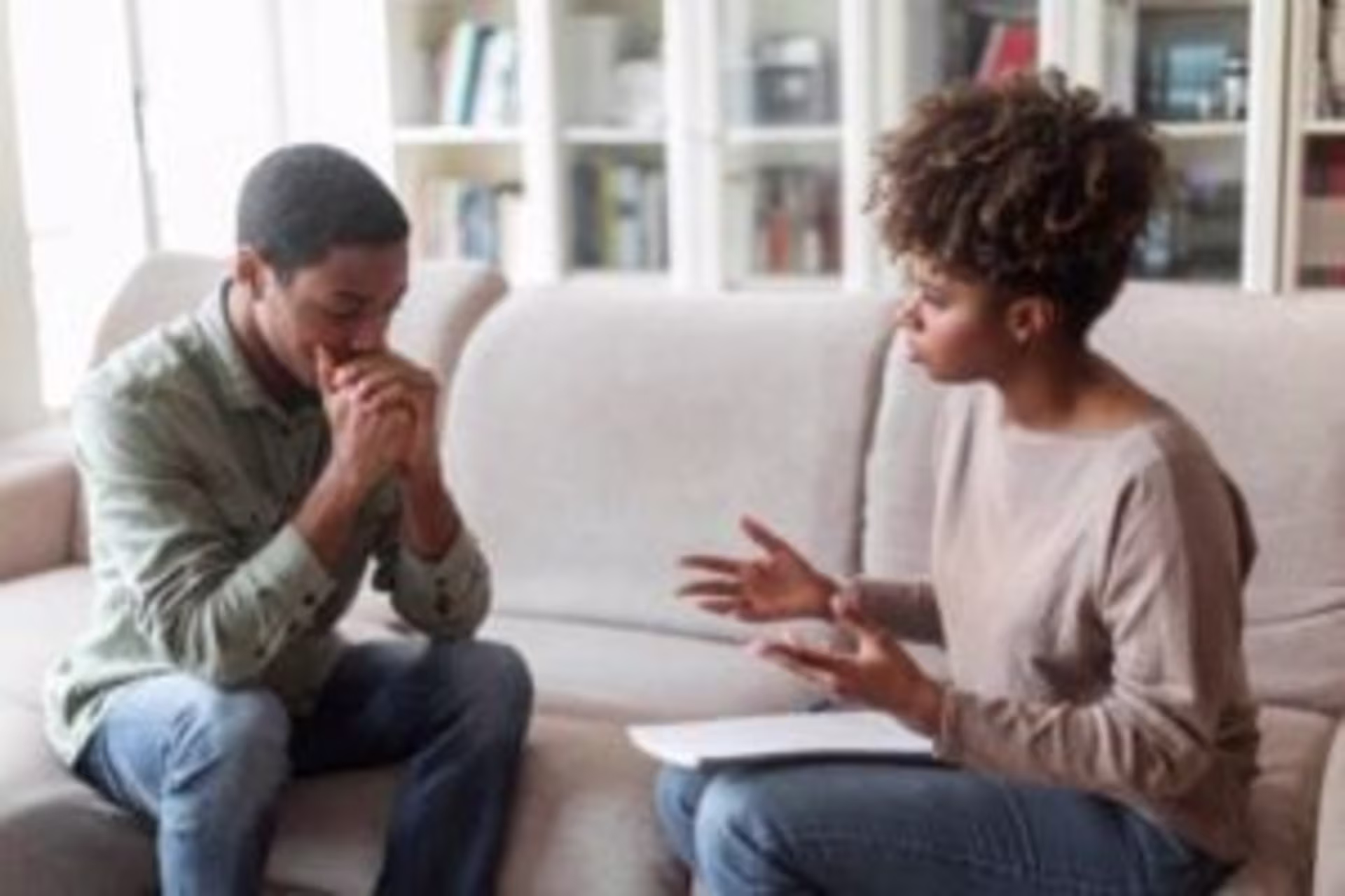 A man and a woman sit on a couch in San Francisco, engaged in a serious conversation. The woman, likely the right San Francisco therapist, holds a notepad while the man appears contemplative with his hands clasped. Bookshelves are in the background.