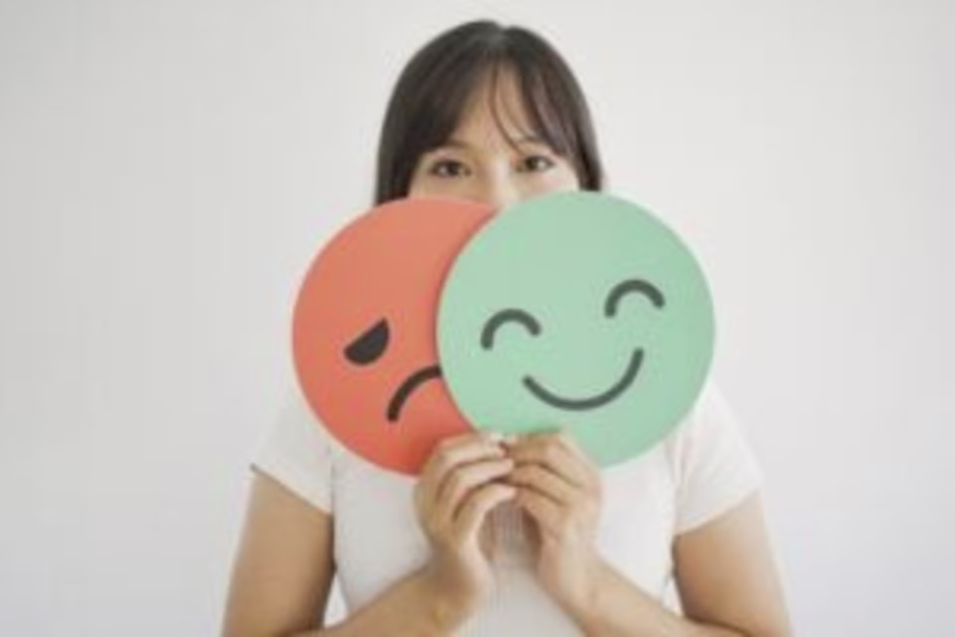 A woman holds a sad red face and a happy green face sign in front of her, partially covering her own face against a plain background, symbolizing the struggles of ADHD masking and revealing true emotions.