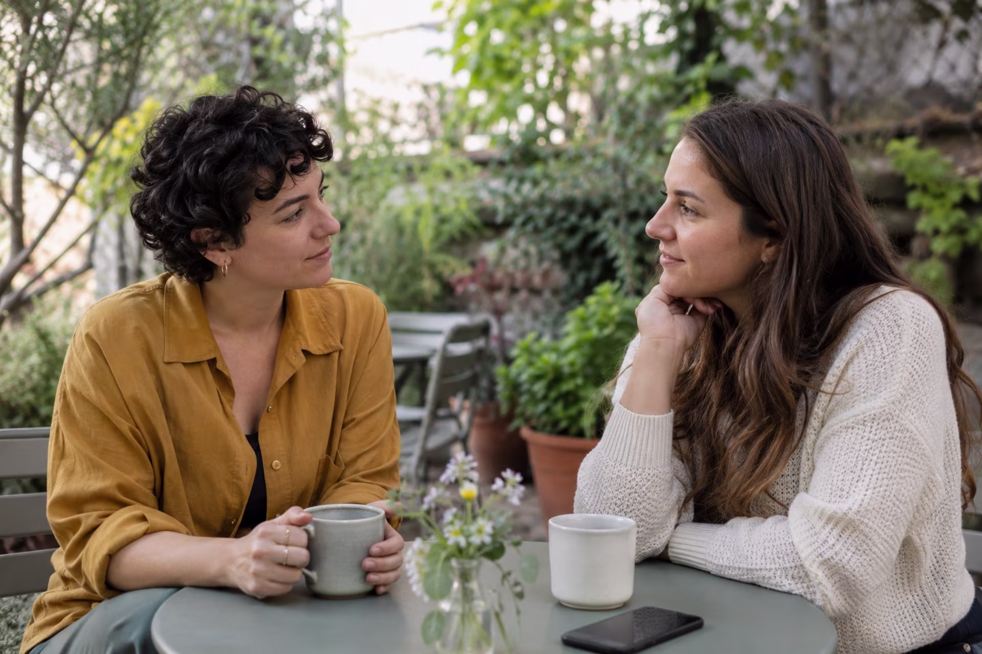 Two women talk over coffee at an outdoor garden table.