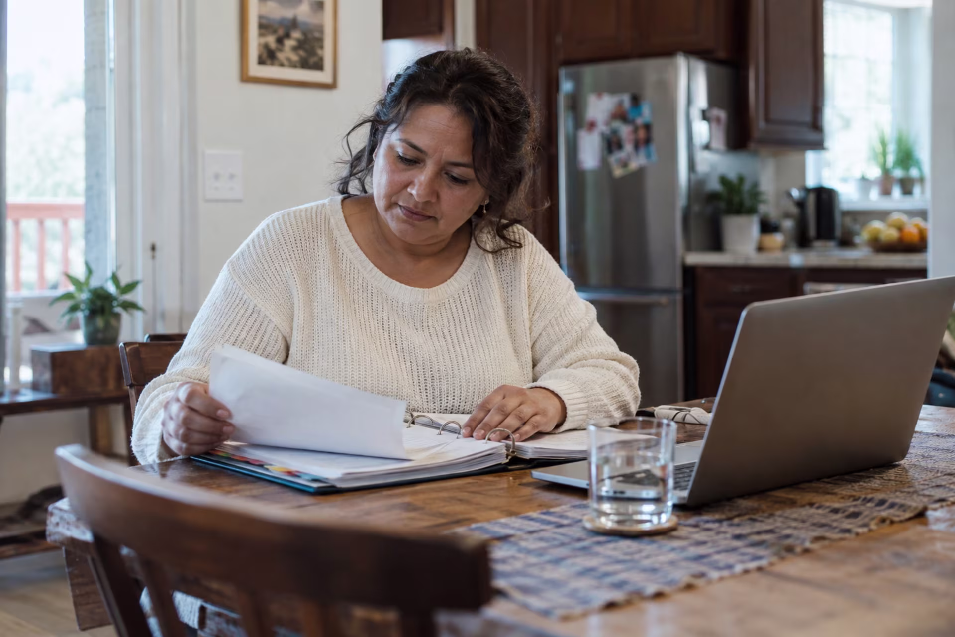 A woman reviews papers in a binder at her kitchen table next to a laptop, organizing documents for an ADHD disability application.