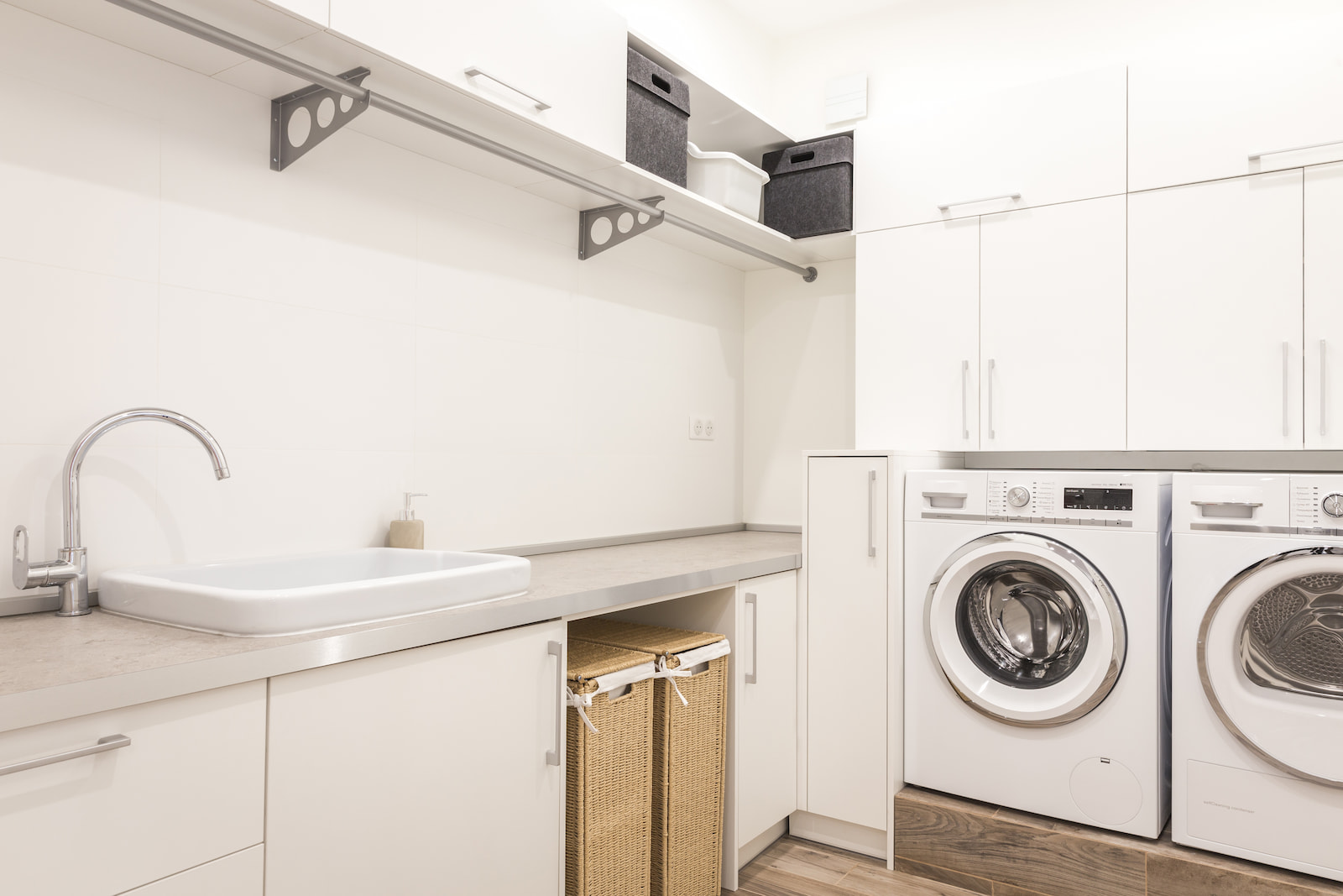Modern laundry room with white cabinetry, front-load washer and dryer, utility sink, and storage baskets.
