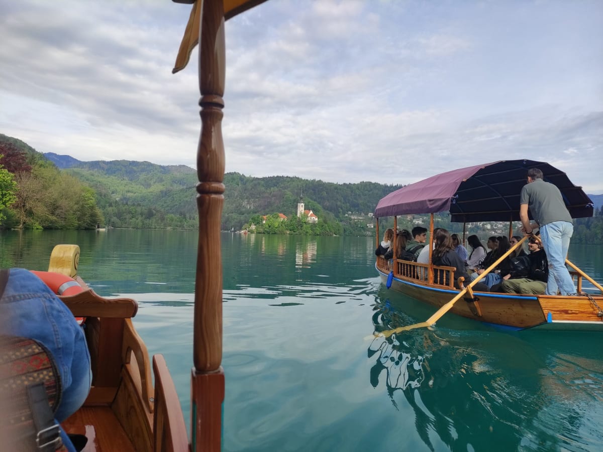 Looking past a carved wooden post from a pletna boat toward a second boat full of passengers, the island church on its green islet far ahead across turquoise water