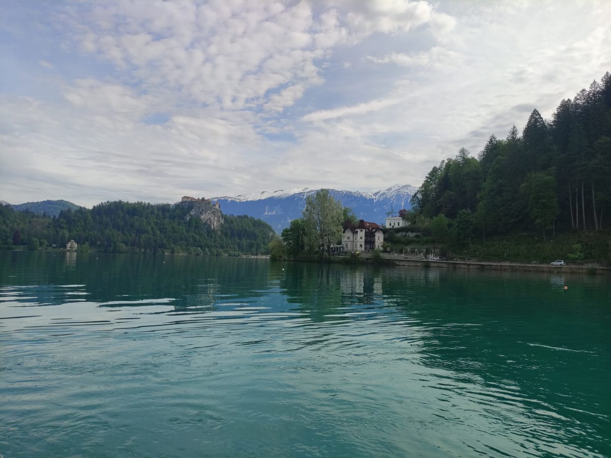 The wooded shore of Lake Bled from the water, a villa among the trees and snow-capped peaks showing under a layered sky