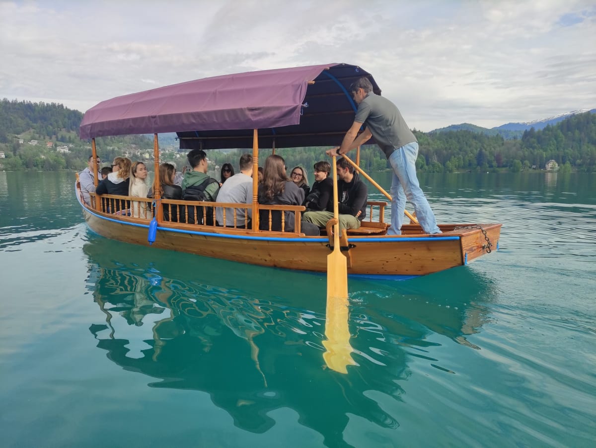 A covered wooden pletna boat full of passengers on Lake Bled, the boatman standing at the stern working a single oar, wooded shore and hazy hills behind