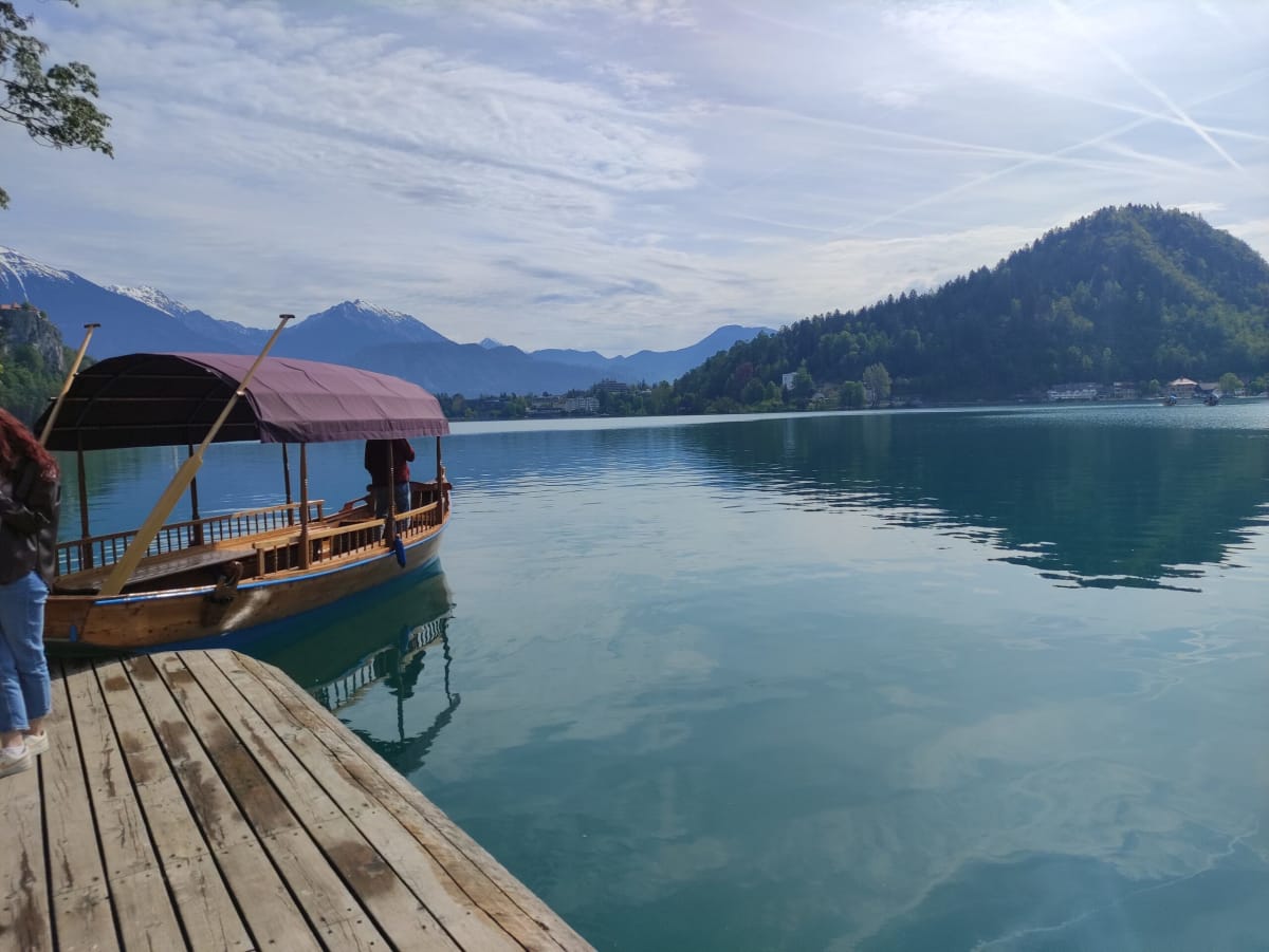 A single pletna boat moored at the island jetty with its oars upright, the lake opening out toward Osojnica hill and hazy Alps