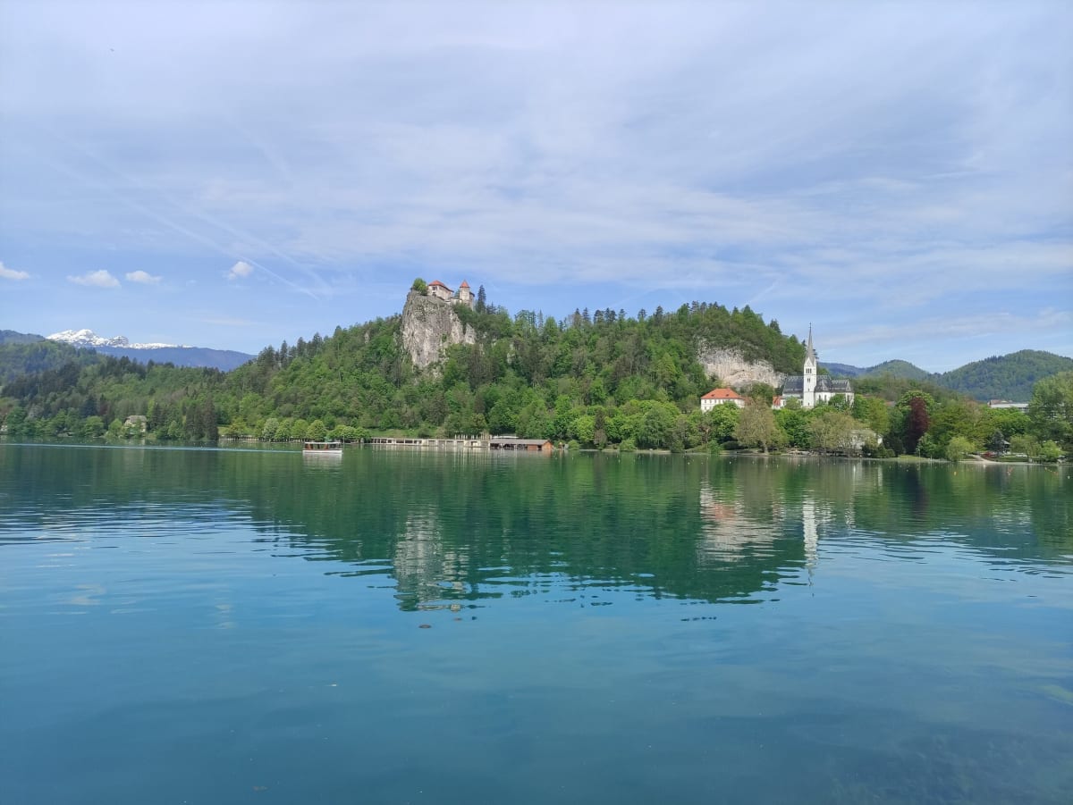 The classic Lake Bled view — the clifftop castle and the neo-Gothic church of St Martin on the wooded shore, reflected in still green water, snowy peaks to the left
