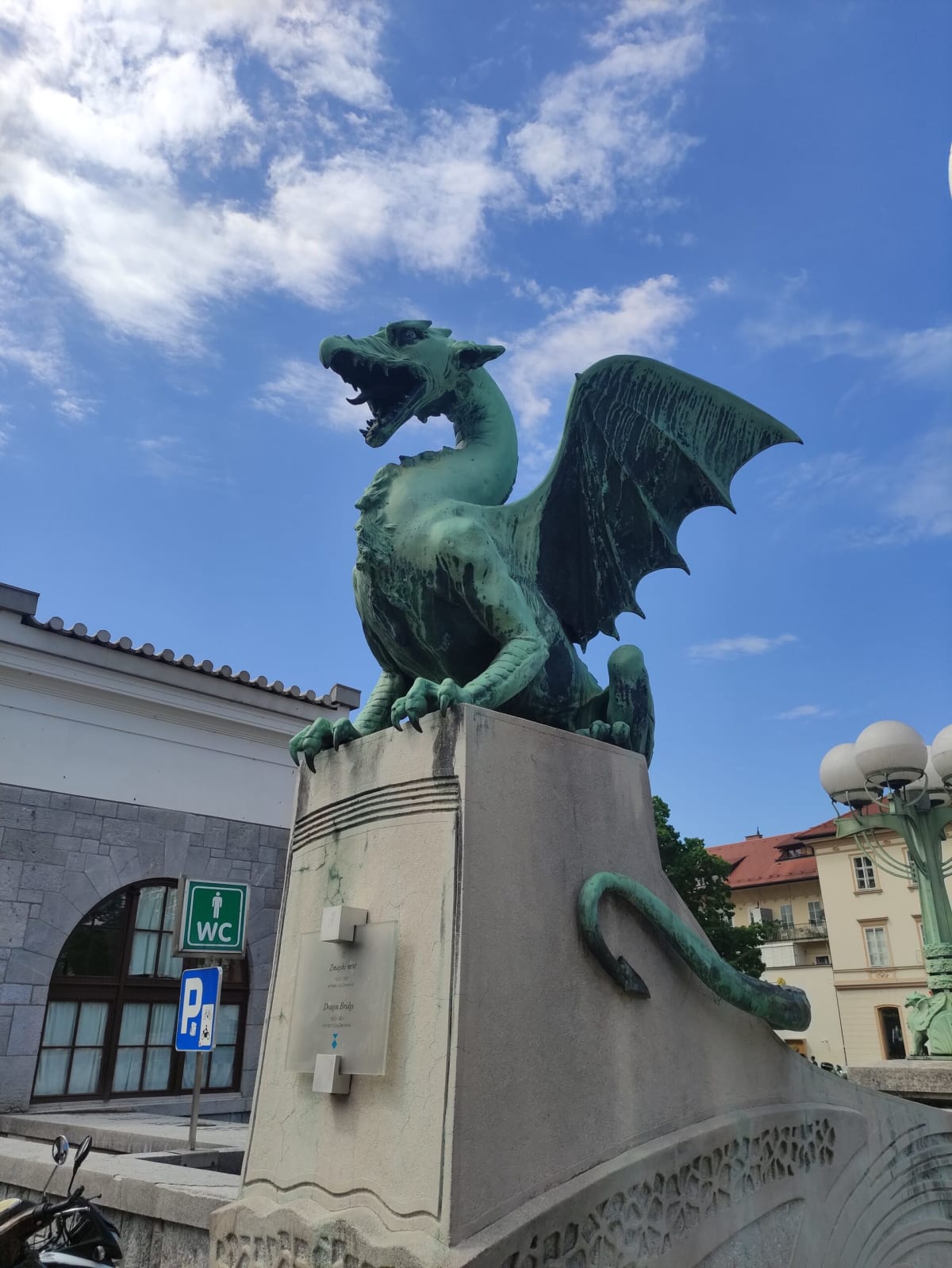 A green bronze winged dragon on Ljubljana's Dragon Bridge, mouth open against a blue sky, Art Nouveau lamp-posts behind