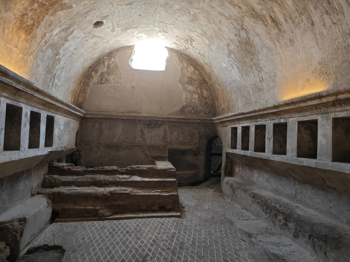 The vaulted interior of a Pompeii bathhouse room, a skylight in the dome and recessed niches along the walls above a stepped floor