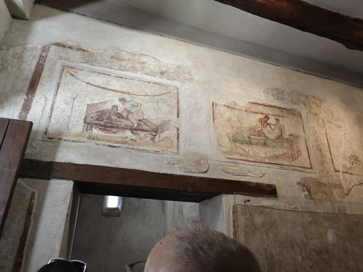 Faded erotic frescoes in small panels above a doorway in a Pompeii building, a visitor's head in the foreground