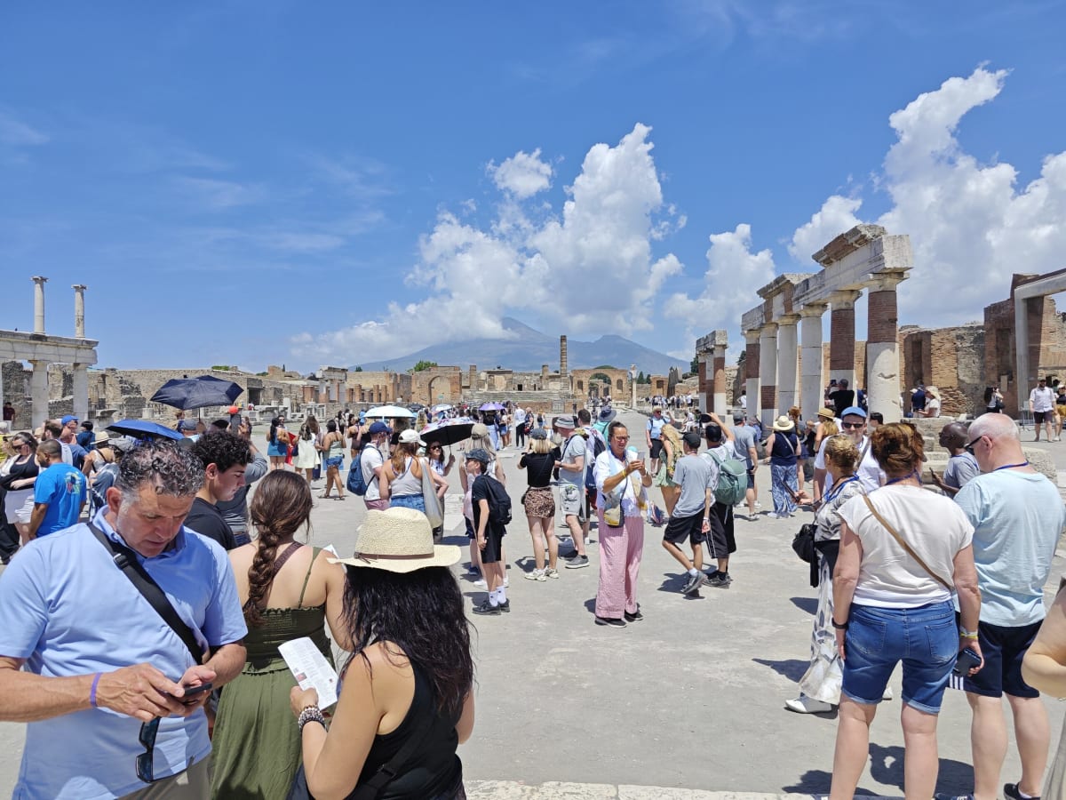 The crowded Forum of Pompeii under a bright sky, visitors with umbrellas and a line of columns, Vesuvius faint on the horizon
