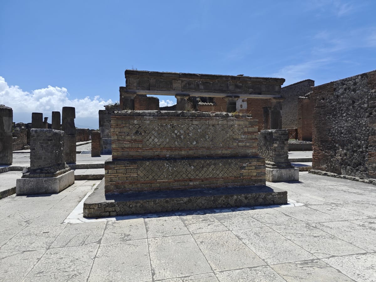 A brick-and-stone podium among the ruins of the Forum, columns and walls beyond and clouds building on the horizon