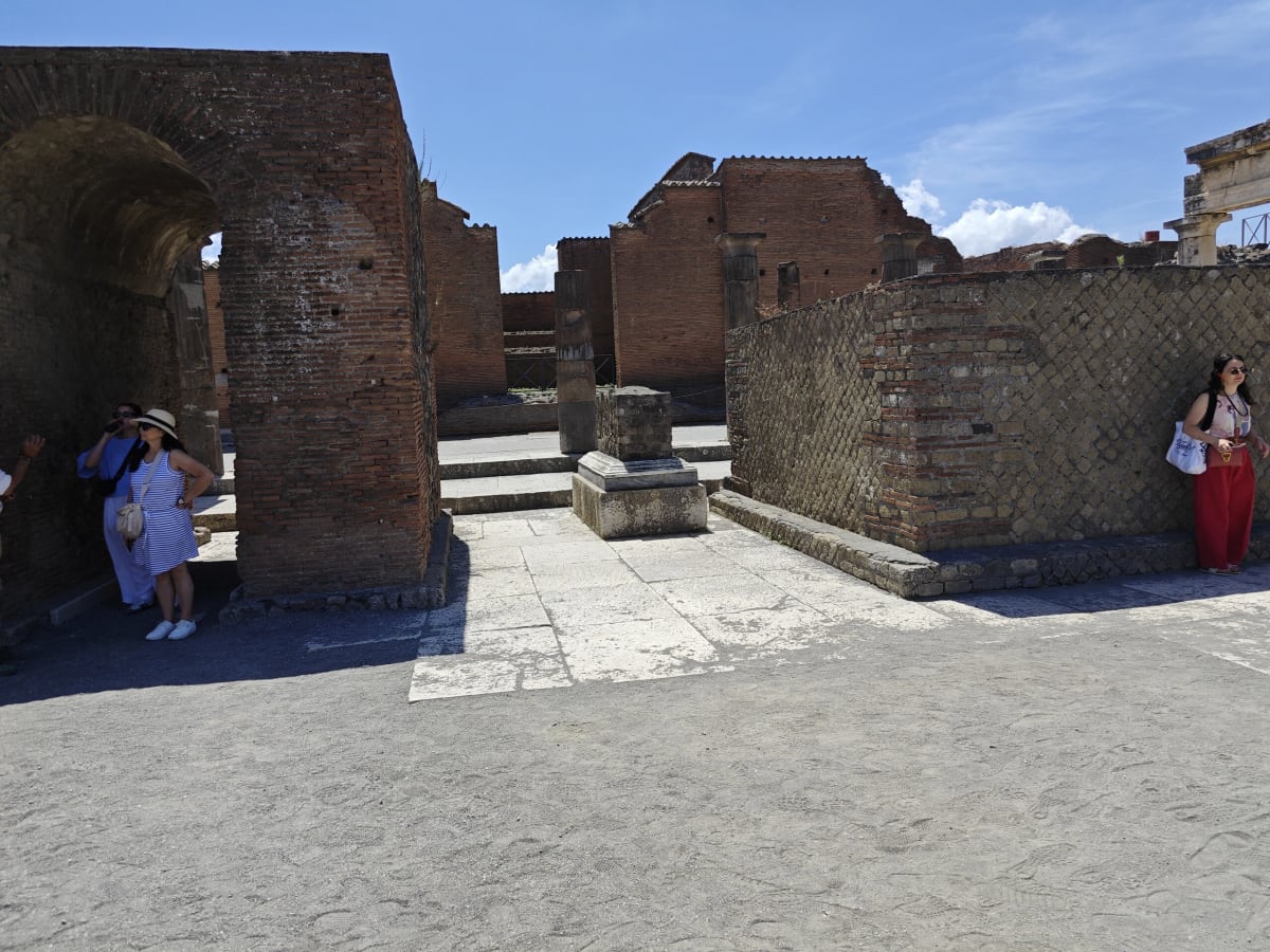 A tall brick archway and a low latticed brick wall in the Forum area, a visitor sheltering in the shade of the arch