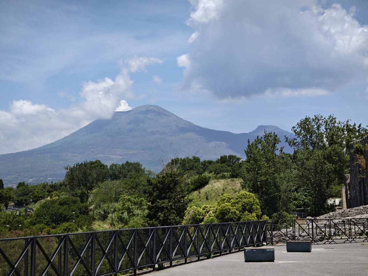 Mount Vesuvius rising behind the trees beyond Pompeii, seen from a railed terrace with clouds gathering at the summit