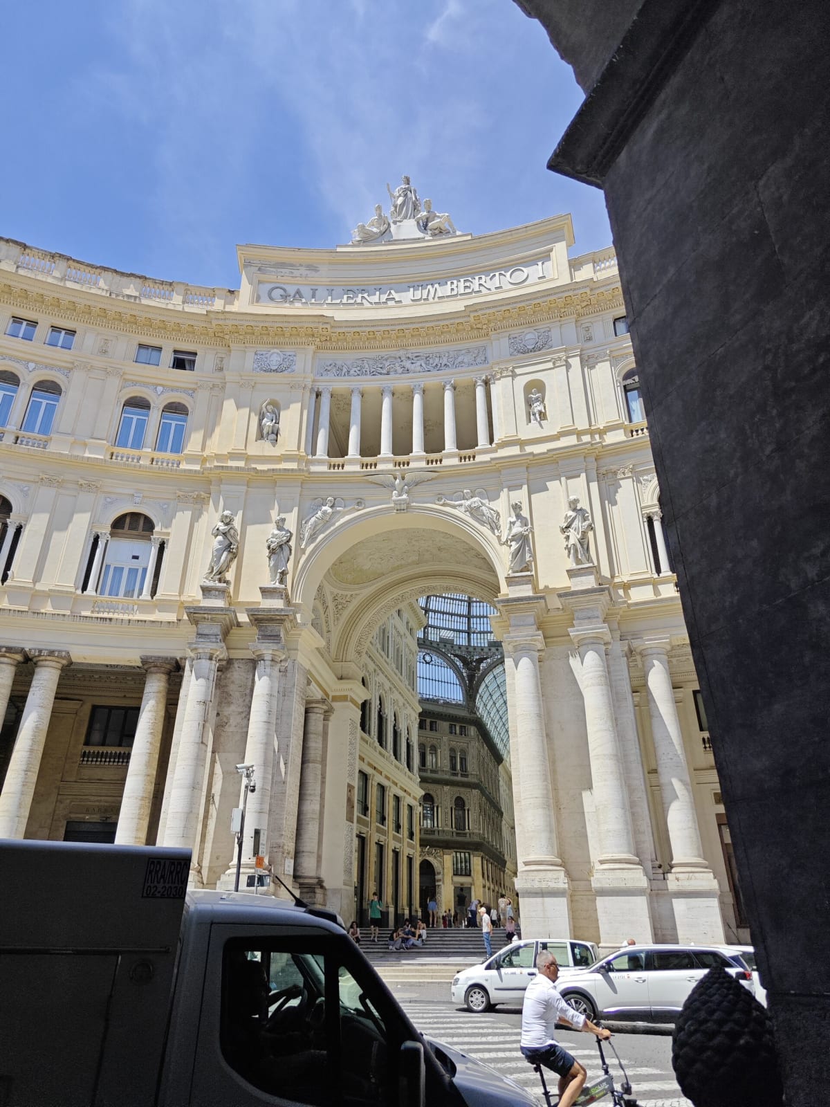 A curved Neapolitan arcade with a cream-coloured balustraded façade and the lettering Galleria Umberto I, seen across a sunlit street