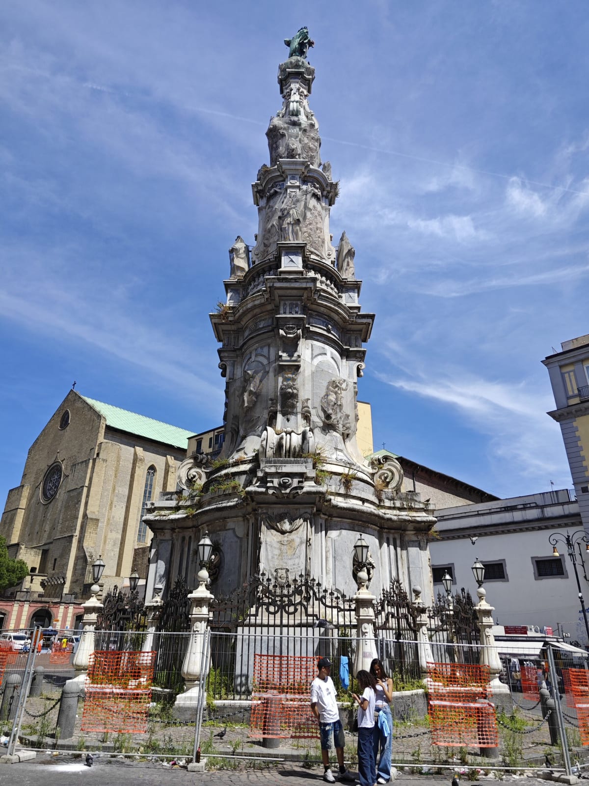 The Guglia dell'Immacolata in Naples — a tall, ornate baroque obelisk of carved stone covered in statues, behind orange site fencing