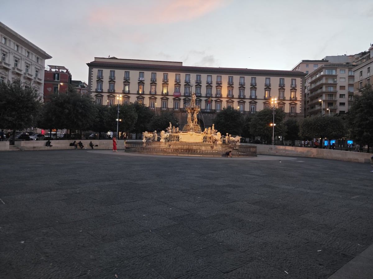 A wide dark Neapolitan square at dusk with a lit fountain in the middle and a long palazzo with glowing windows behind
