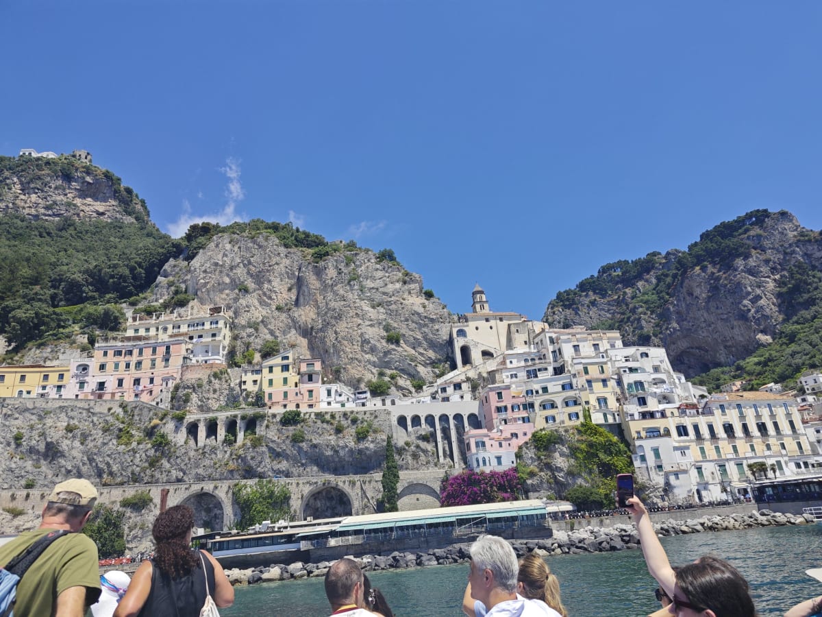 Amalfi from the water — pastel houses stacked up a cliff beneath rocky peaks, the dome of the cathedral at the centre, passengers along the rail