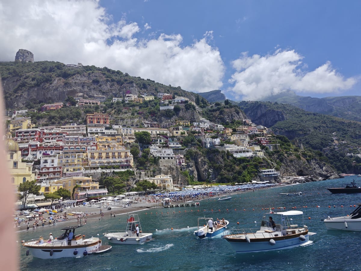 Positano from the sea, colourful houses climbing the hill above a pebble beach, small boats and motor launches in the foreground
