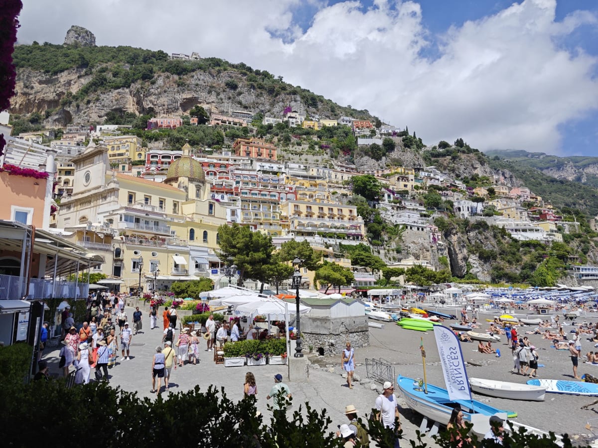 The promenade at Positano — a crowd on the waterfront under the church dome and the steep terraced hillside, beach umbrellas at right
