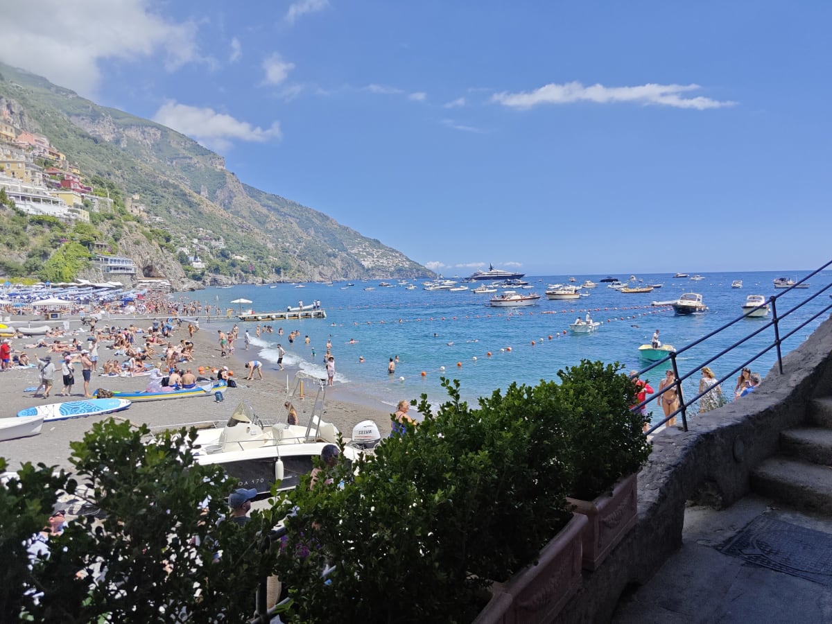 The beach at Positano seen from a stone stairway, sunbathers on the sand and boats floating in turquoise water below the cliff