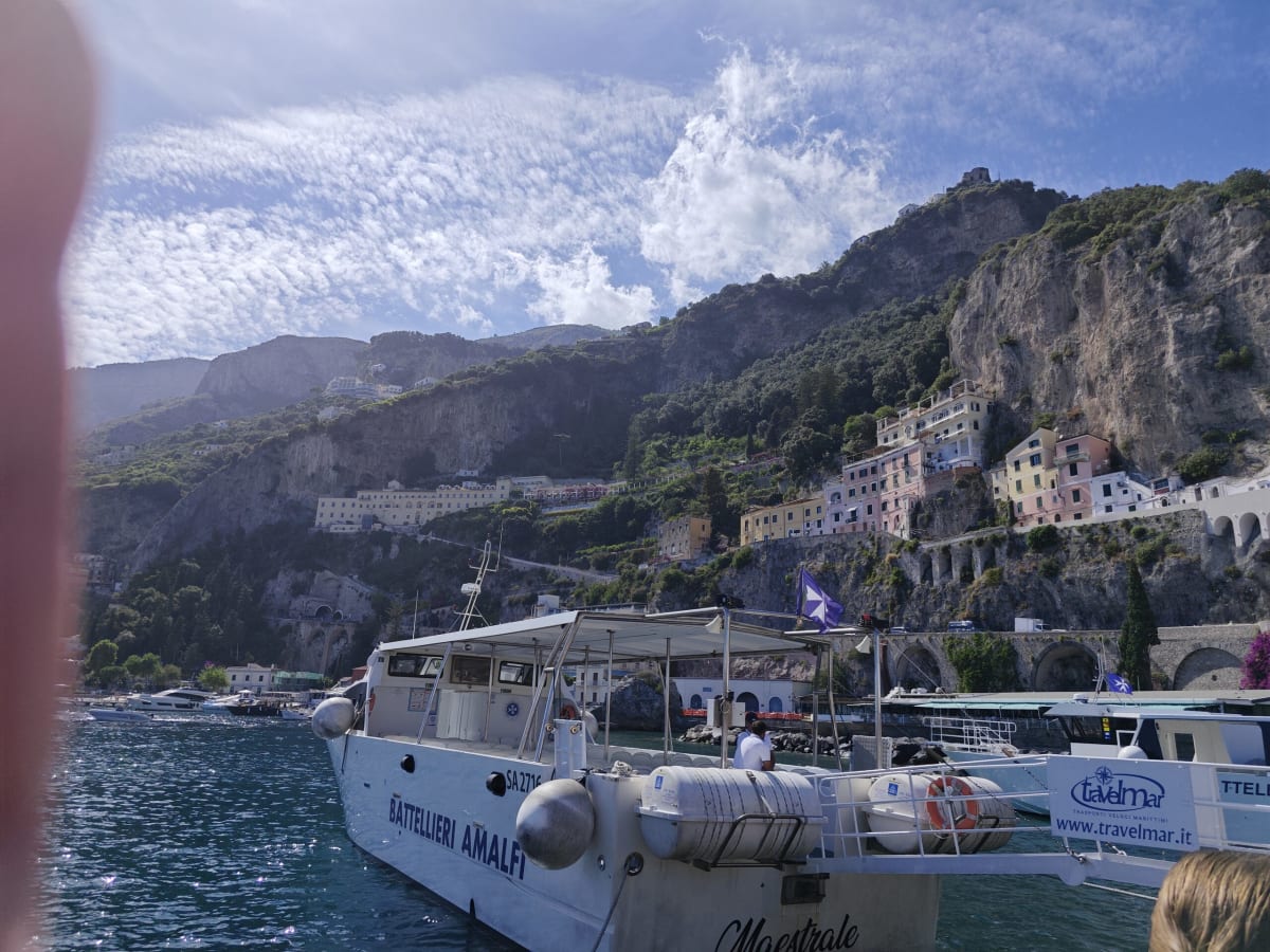 An Amalfi ferry boat moored below a cliff of pastel houses and tunnel arches, thin clouds streaking the sky