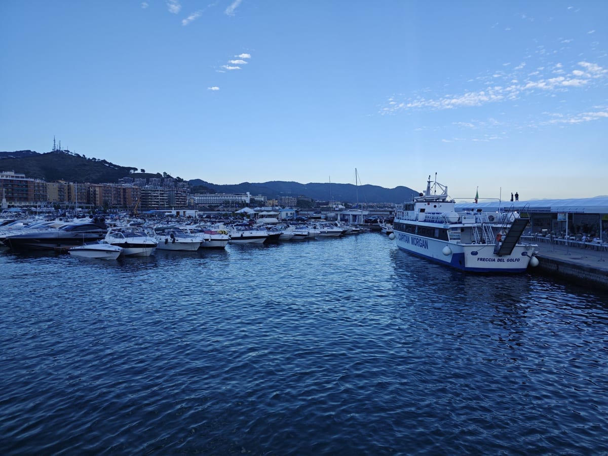 A marina in the evening light, a large white ferry at the quay and small boats moored in dark blue water under hazy hills