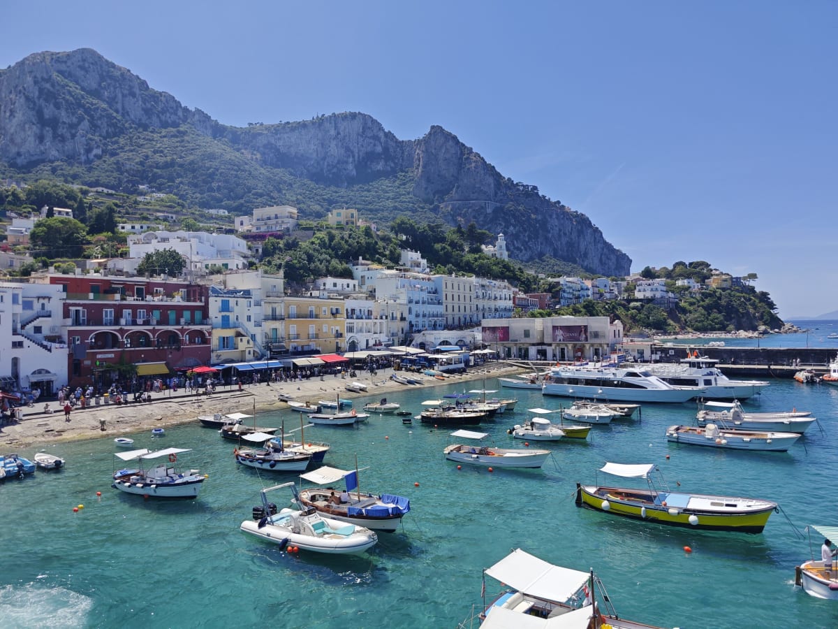 Capri's Marina Grande from above, a ring of pastel buildings at the foot of a limestone cliff, small boats in turquoise water