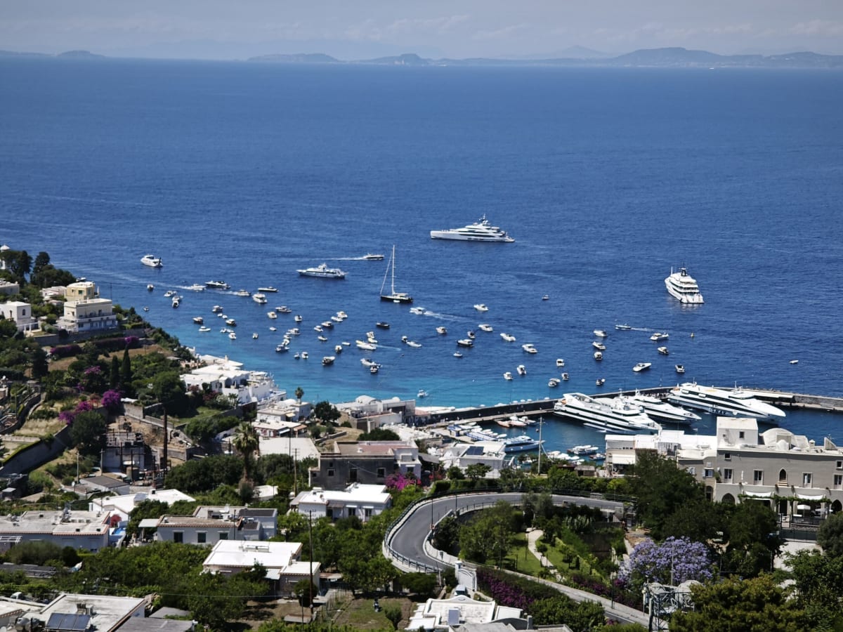 Looking down on a hairpin road and white villas above the bay of Capri, yachts anchored in dark blue water and distant islands on the horizon