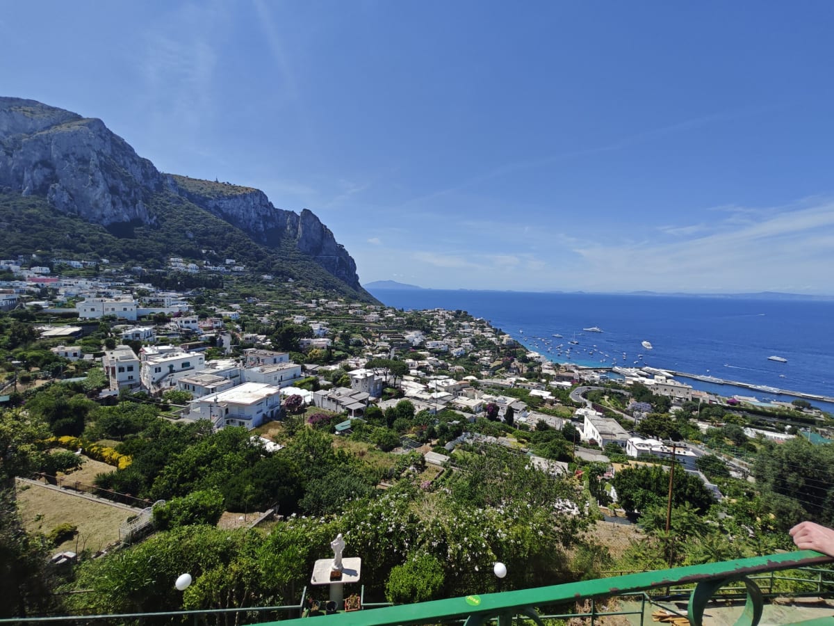 A view from above Capri over green hillsides and white houses to the sea, a cliff rising at left