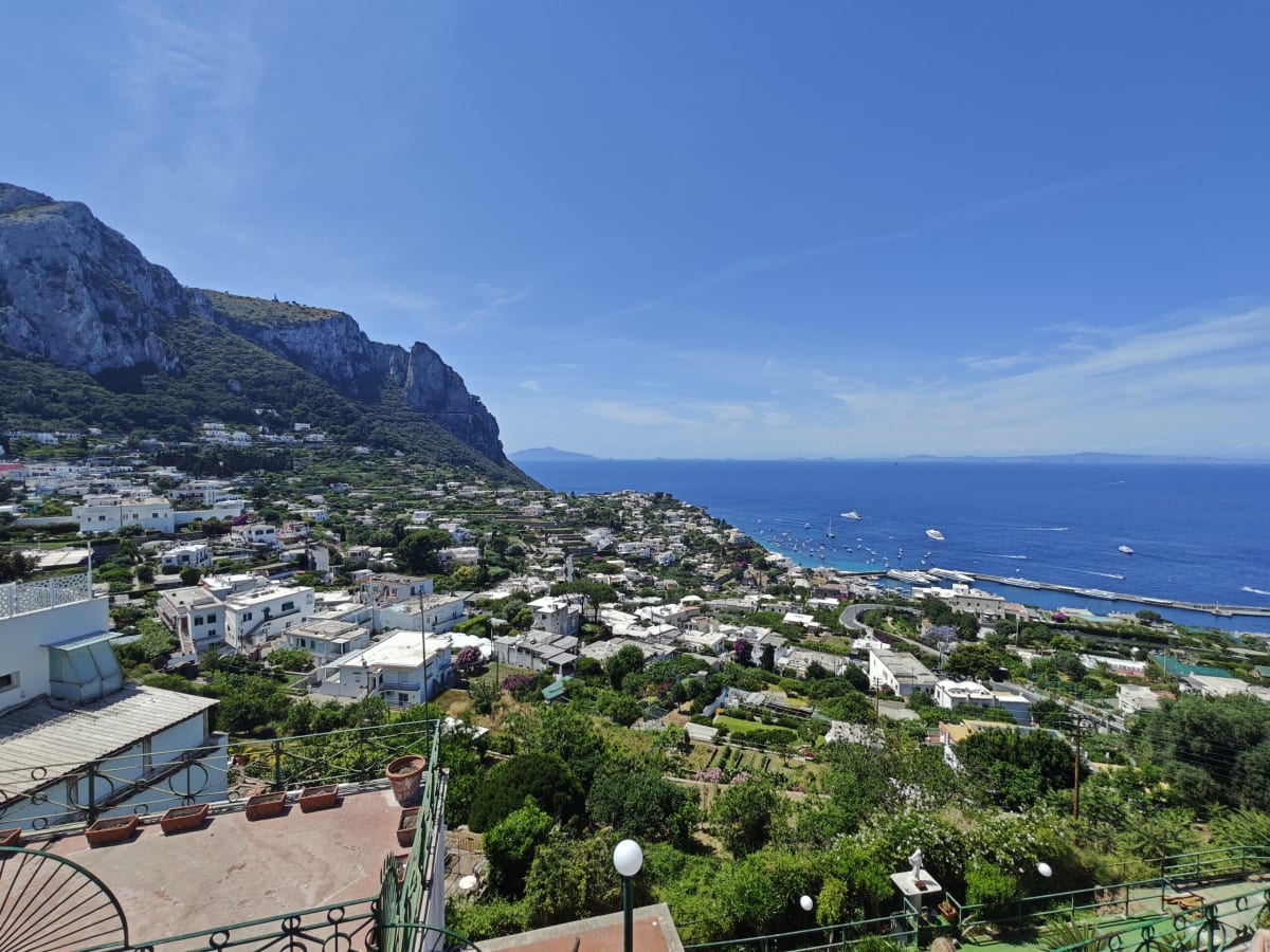 The same Capri view from a terrace with tiled floor and lamps in the foreground, the headland, harbour mole and sea beyond