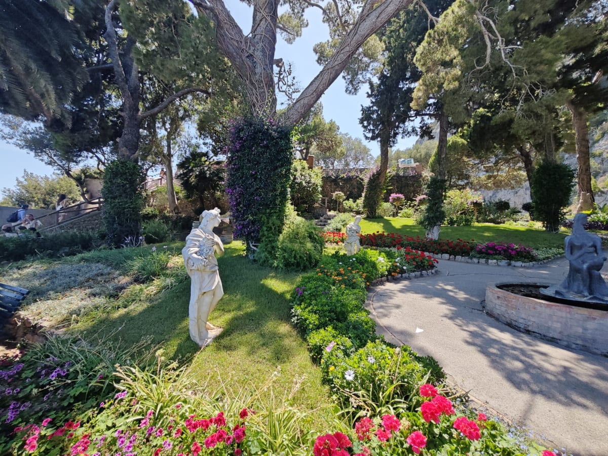 A walled flower garden on Capri — a white marble statue on a lawn, red geraniums and a curving gravel path beneath umbrella pines