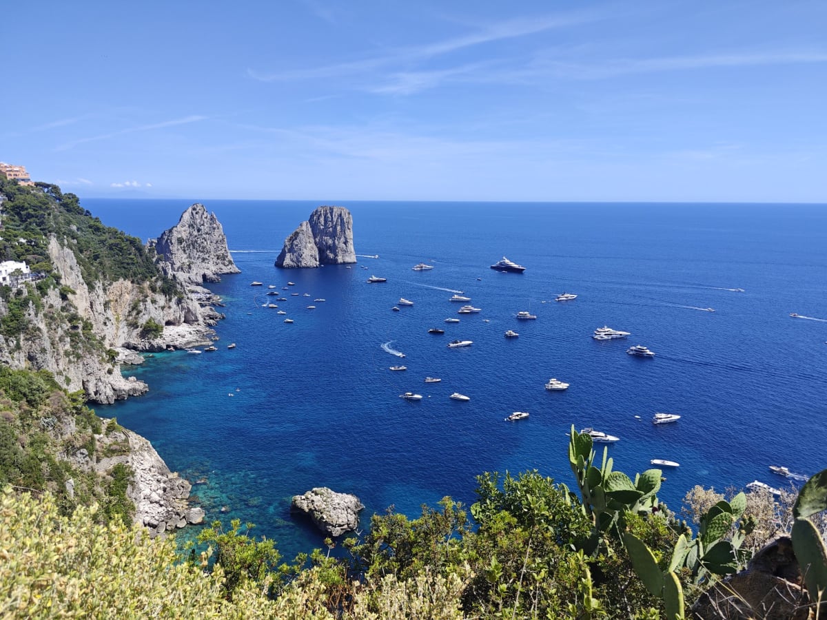 The Faraglioni rock stacks rising from a deep-blue bay, yachts spread across the water below a rugged green slope