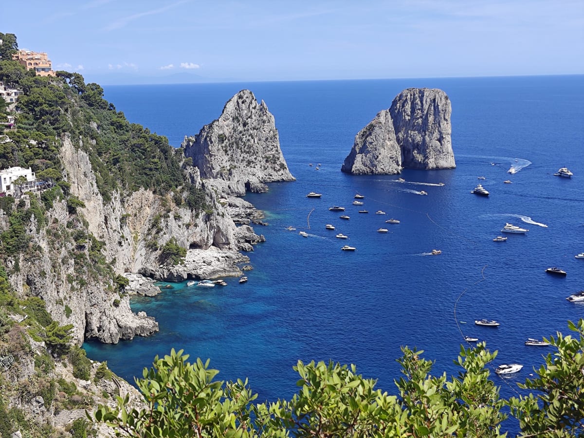 The two Faraglioni pinnacles rising from the blue water off Capri, clear green depths at their foot and boats trailing wakes