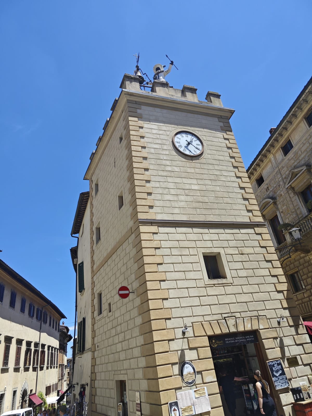 A square stone clock tower with a crenellated top and a weather figure, standing at a corner of a street in a Tuscan hill town