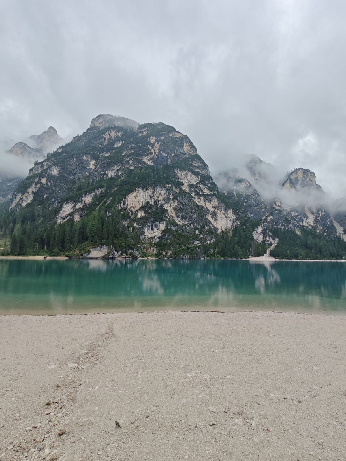 Lago di Braies — turquoise water below the rounded, forested Seekofel cliff, low cloud, an empty gravel shore