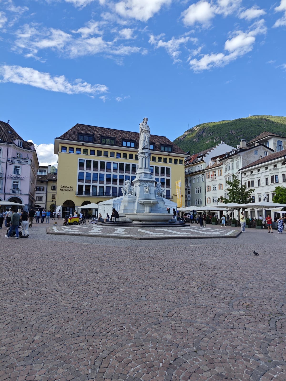 Piazza Walther in Bolzano — the marble Walther von der Vogelweide monument and fountain, café umbrellas, the yellow Cassa di Risparmio building, a green hill behind