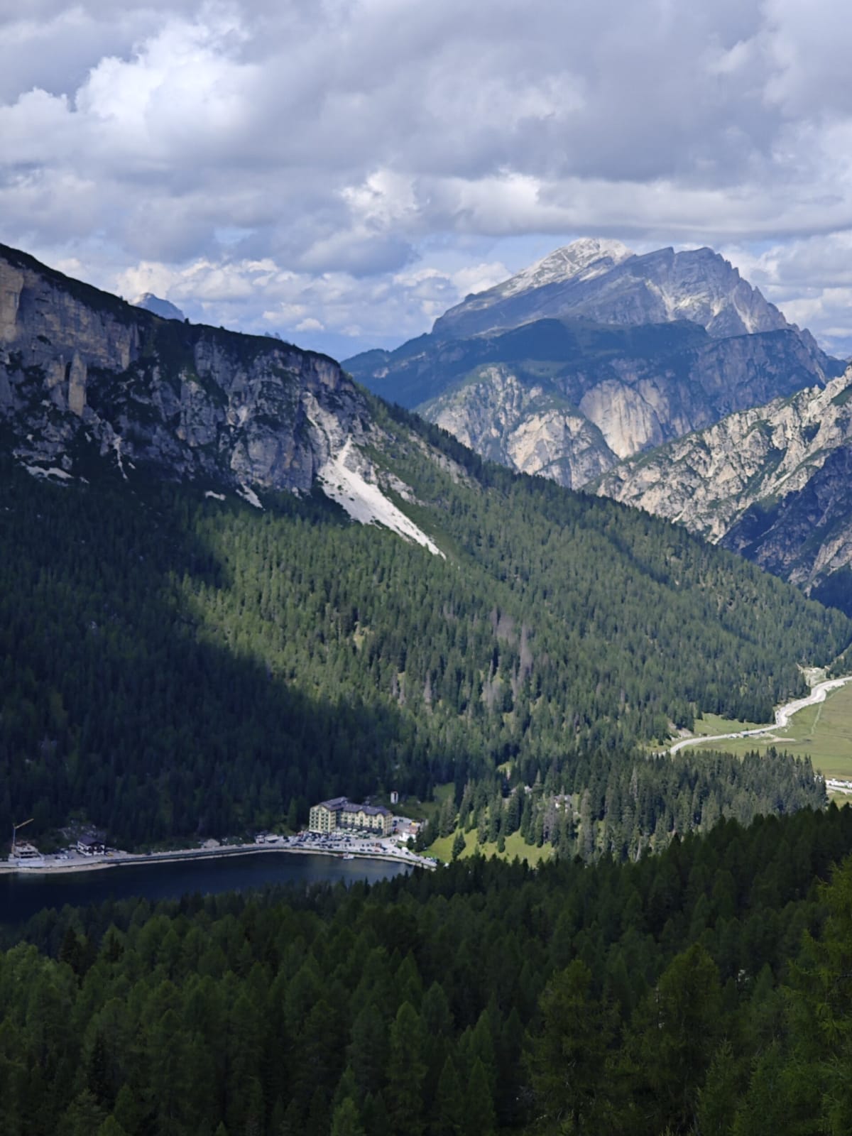 Lago di Misurina and its large lakeside hotel from above, dark conifer forest, the pyramid of Monte Antelao beyond