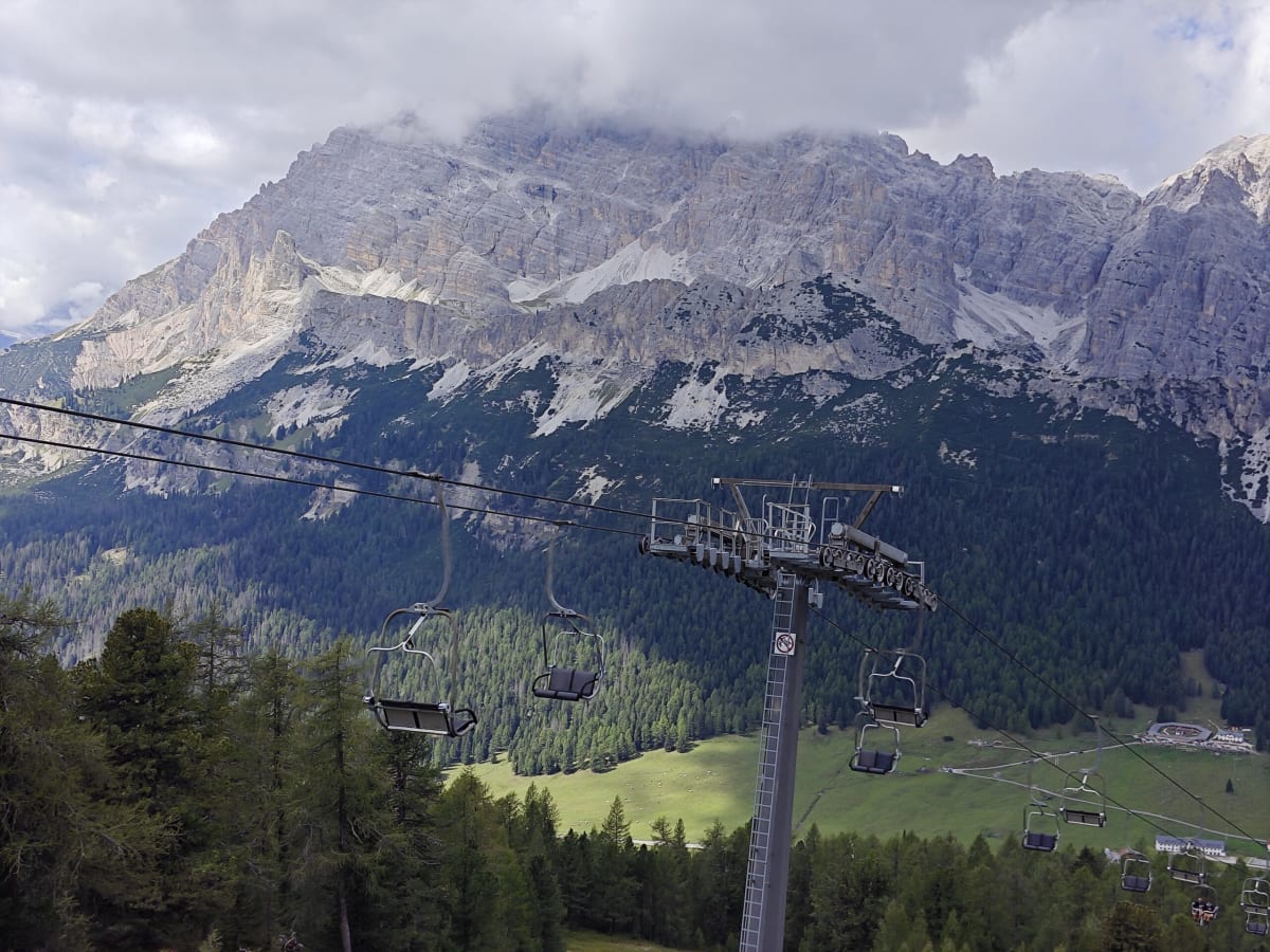 Empty chairlift seats climbing toward the Cristallo wall, green pasture and forest below, cloud on the summit