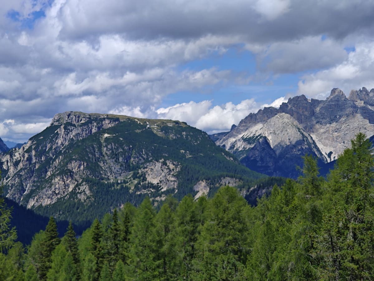 Layered wooded ridges with the spires of the Cadini di Misurina on the right, broken cloud overhead