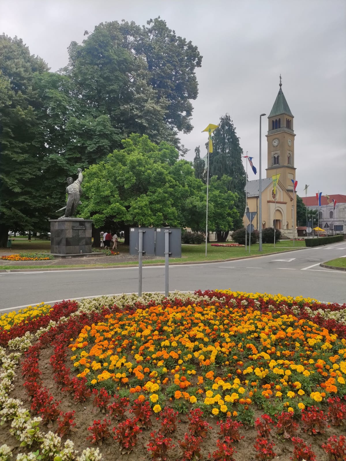 A dense roundabout bed of orange, yellow and red marigolds and begonias in central Đurđevac, a bronze figure on a plinth and the yellow clock tower of St George's church behind under a grey sky