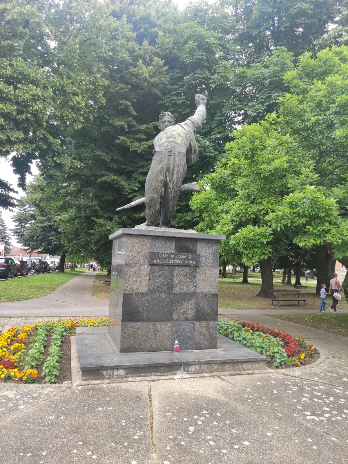 The WWII partisan monument in Đurđevac's town park — a striding bronze figure with a raised fist on a granite plinth reading "Palim borcima u antifašističkoj borbi", flower beds around it